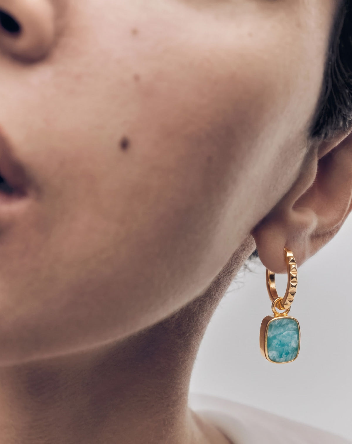 A close-up of a person's ear wearing a gold huggie hoop earring with a rectangular turquoise-blue stone pendant.